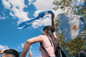 Mujer sostiene una bandera argentina en alto durante una celebración al aire libre bajo un cielo despejado.